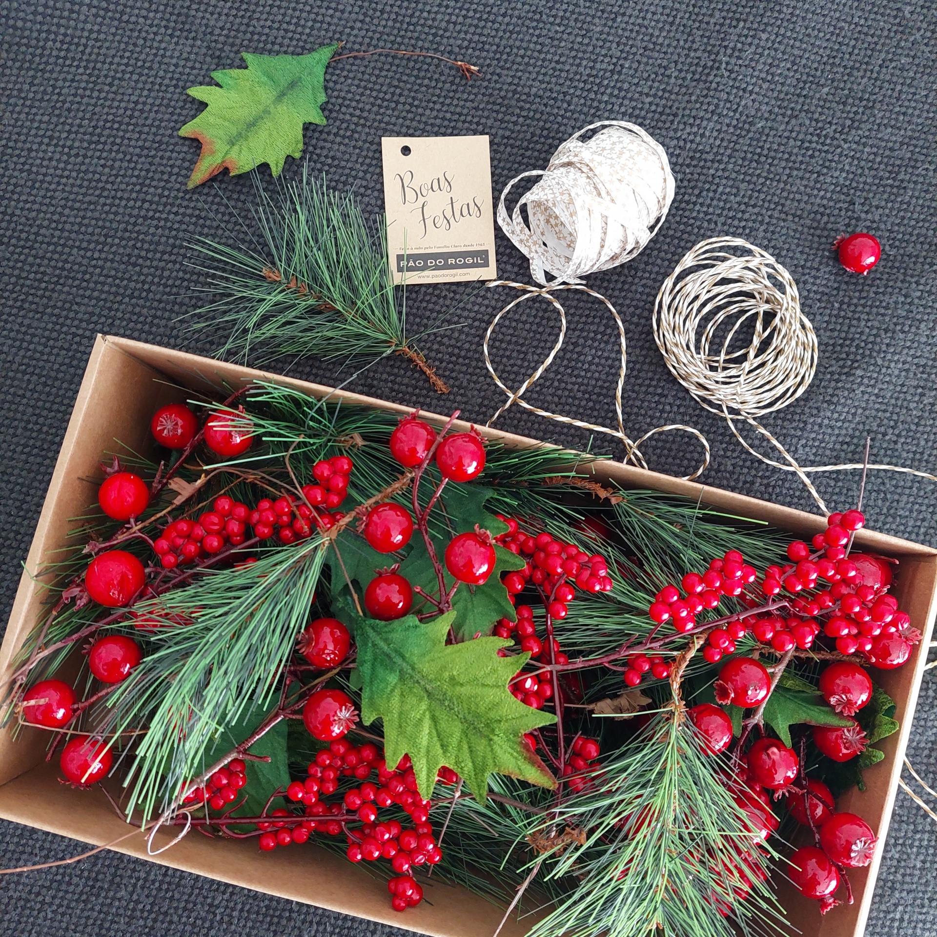 Decoração natalícia com ramos verdes, bagas vermelhas e etiqueta Boas Festas em caixa aberta