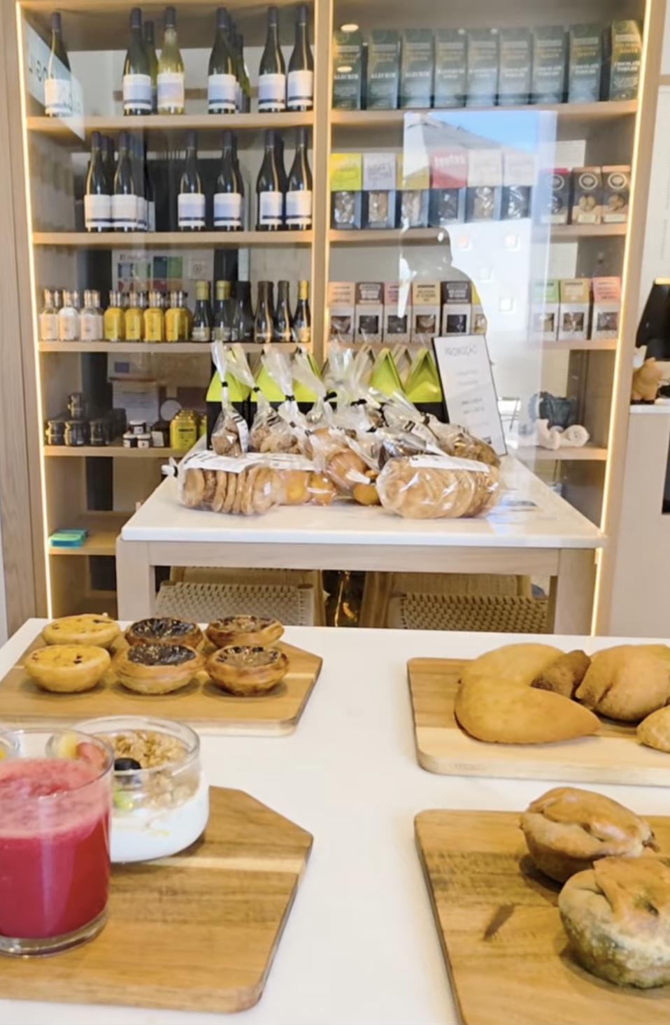 Produtos variados incluindo vinhos, bolachas, pastéis de nata e bebida sobre mesa branca e prateleiras de madeira
