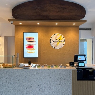 Balcão de mármore com amostras, painel castanho com placa circular da Geladaria PÃO DO ROGIL e caixa registadora digital.