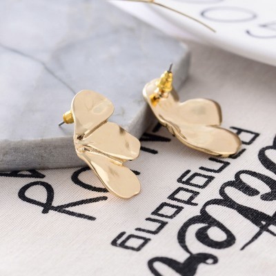 Brincos dourados abstratos em três pétalas sobre mármore cinza e tecido branco com texto