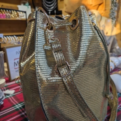 Mala saco dourada texturizada com alça ajustável sobre mesa com toalha xadrez e acessórios