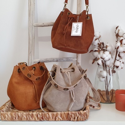 Três bolsas tipo saco em camurça em diferentes tons, sobre bandeja e penduradas numa escada, vaso com flores de algodão e vela.