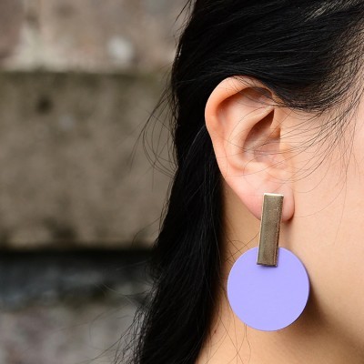 Brinco pendente dourado com disco lilás na orelha de uma pessoa