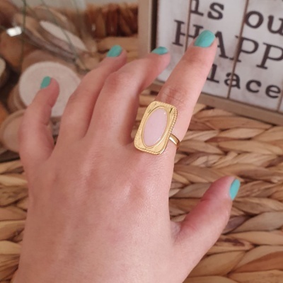 Anel dourado com pedra rosa em mão com unhas azuis sobre superfície de palha