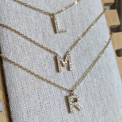 Três colares dourados com letras L, M e R em cristais sobre tecido bege