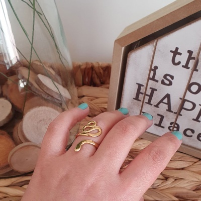 Anel dourado em forma de serpente numa mão com unhas azuis sobre superfície de ráfia e decoração ao fundo