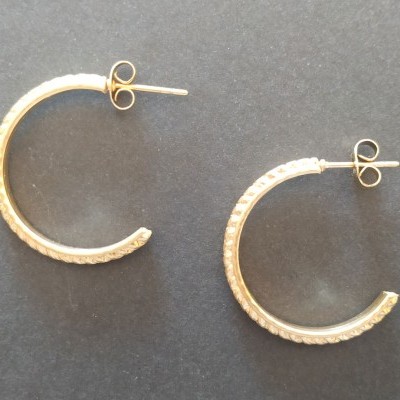 Brincos de argola dourados semiabertos com textura sobre fundo cinzento