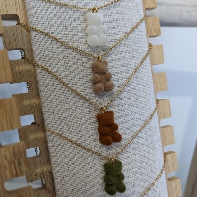 Colares com pendentes em forma de ursinho de pelúcia em várias cores, com correntes douradas, expostos numa base de tecido bege e madeira.