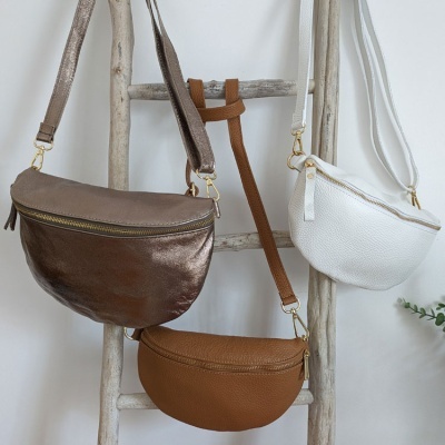 Três malas tipo pochete em prata, branca e castanha penduradas num cabide de madeira.