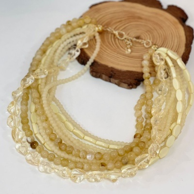 Colar multi-fios bege e branco com contas diversas e fecho dourado sobre base de madeira.