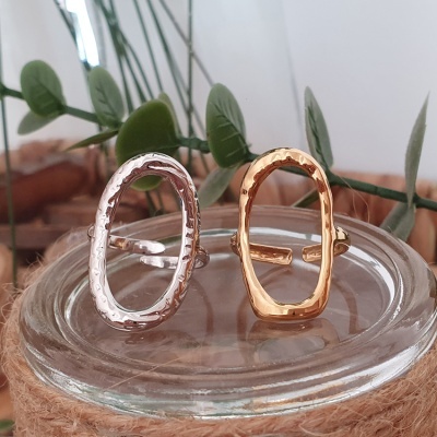Dois anéis ovais, um prateado e um dourado com textura martelada, sobre superfície de vidro com folhas verdes ao fundo