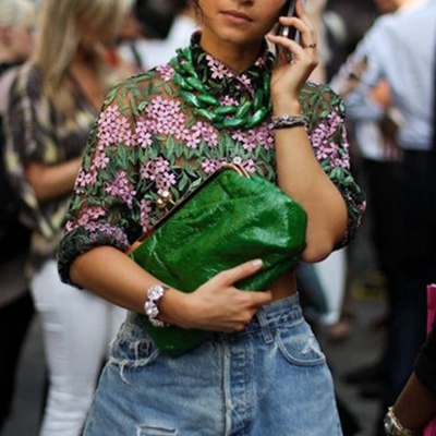 Mulher segura mala verde de pele com blusa floral transparente e calças de ganga