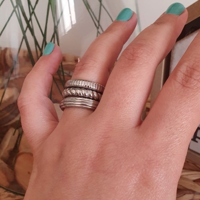 Mão com quatro anéis prateados em dedo, unhas verdes, fundo com objetos e plantas desfocados.