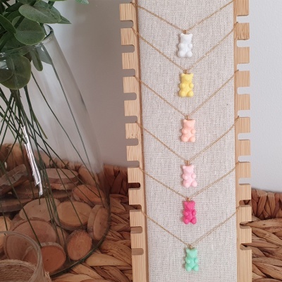 Display com cinco colares de urso em diferentes cores com fundo de tecido bege e vaso transparente com folhas verdes.