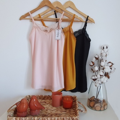 Três vestidos suspensos: rosa claro, mostarda e preto com detalhes de renda, pendurados em cabides de madeira junto a uma parede branca.