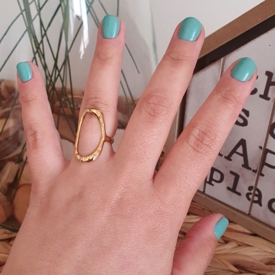 Anel dourado oval em mão com unhas azuis, fundo com decoração e plantas.