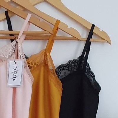Três vestidos ou tops com alças finas pendurados, cores rosa, mostarda e preto, com renda e etiqueta