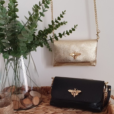 Dois bolsos de mão em pele dourado e preto com detalhe de abelha e corrente dourada, junto a vaso com planta