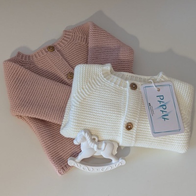 Dois casacos de malha para bebê, rosa e branco, com botões de madeira e etiqueta PAPAIA junto a um cavalo de baloiço branco.