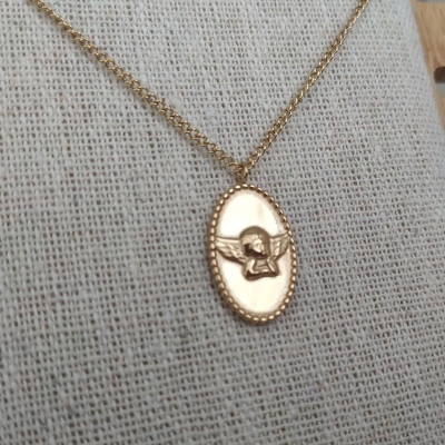 Colar dourado com pendente oval com anjo em relevo sobre tecido bege.
