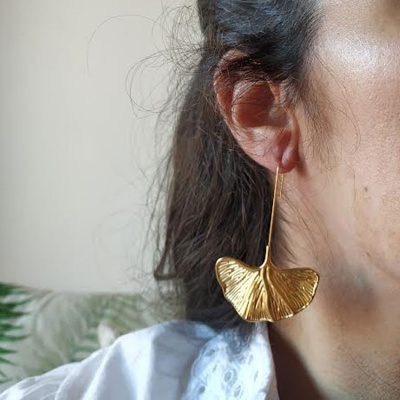 Brinco pendente dourado em forma de folha com textura detalhada, visto de perto no ouvido de uma pessoa