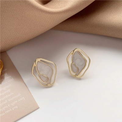 Brincos dourados com centro branco translúcido e textura marmoreada sobre superfície branca com tecido bege ao fundo