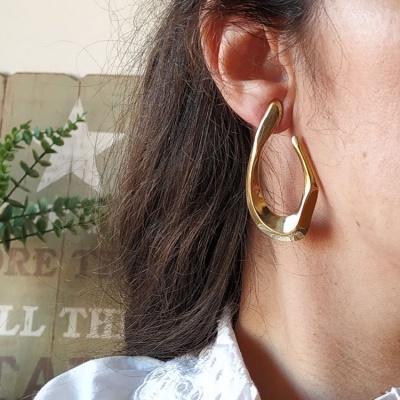 Brinco dourado pendente, forma irregular, orelha direita, fundo almofada bege com texto.