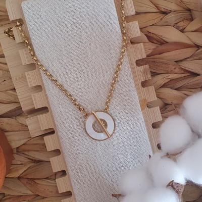 Colar dourado com pendente redondo branco sobre tecido claro e base de madeira