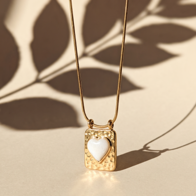 Colar dourado com pendente retangular com coração branco e sombra de folhas ao fundo.