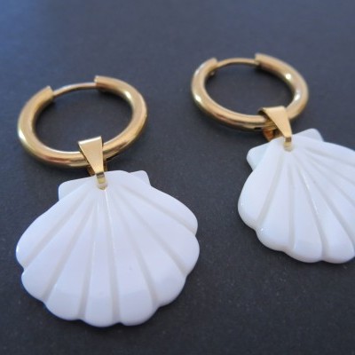 Brincos de argola dourada com pendente de concha branca sobre fundo preto