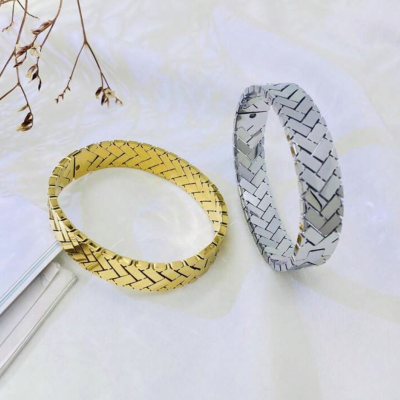 Pulseiras metálicas dourada e prateada com padrão preto, sobre tecido branco ao lado de ramo seco e livro