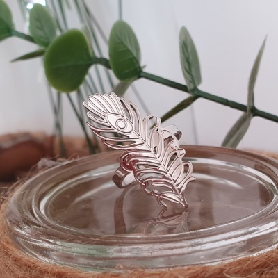 Anel prateado em formato de pena sobre recipiente de vidro com folhas ao fundo