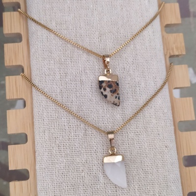 Dois colares dourados com pendentes em padrão animal e branco exibidos em suporte de tecido