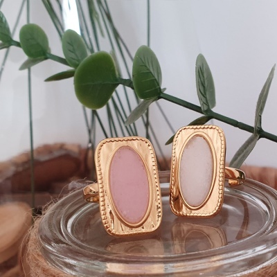 Dois anéis dourados com pedras ovais rosa e branca sobre suporte de vidro