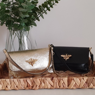 Dois sacos de mão, dourado e preto, com abelha dourada, em tabuleiro de vime com jarra e folhas verdes