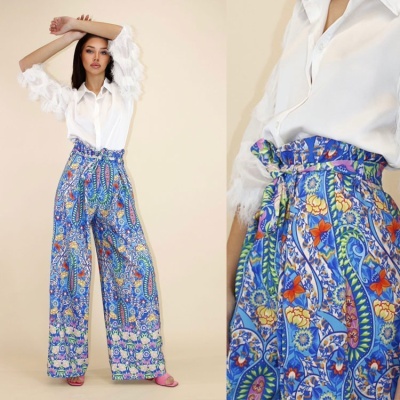 Calças largas de padrão floral colorido com cintura alta e cinto, usadas com camisa branca de mangas compridas com detalhes transparentes.