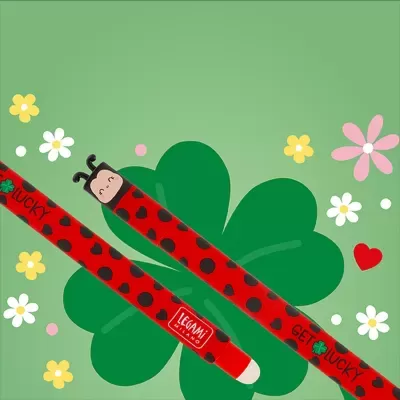 Lápis vermelho com decoração de corações pretos e rosto de joaninha na capa, sobre fundo verde com trevo e flores