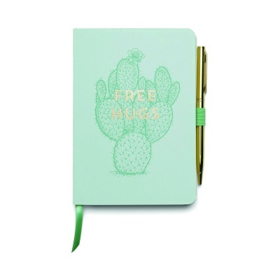 Caderno verde claro com desenho de cacto e texto FREE HUGS e caneta dourada ao lado