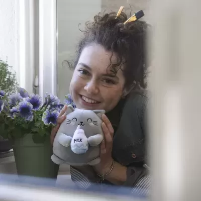 Almofada de gato cinzenta segurando garrafa com texto MILK, com vaso de flores ao lado