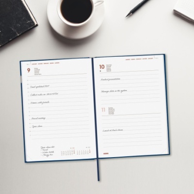 Agenda aberta com anotações, chávena de café, caneta e caderno preto sobre mesa branca