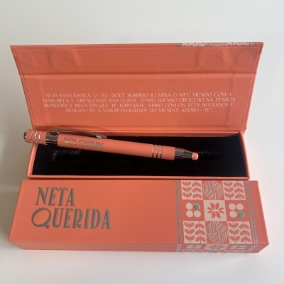 Caneta coral com inscrição NETA QUERIDA numa caixa coral com texto em português e padrão de flores
