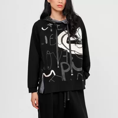 Mulher com sweatshirt preta de capuz estampada com letras e formas abstratas, calças pretas largas, fundo branco.