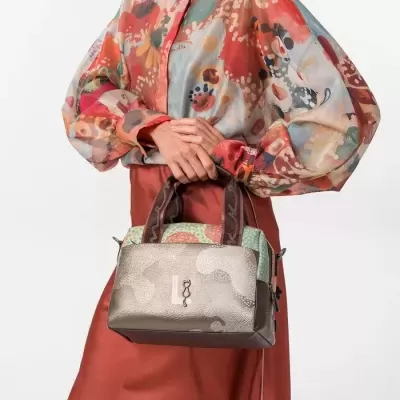 Mala de mão castanha com padrão texturizado e alças castanhas, segurada por uma mulher com blusa colorida e saia castanha.