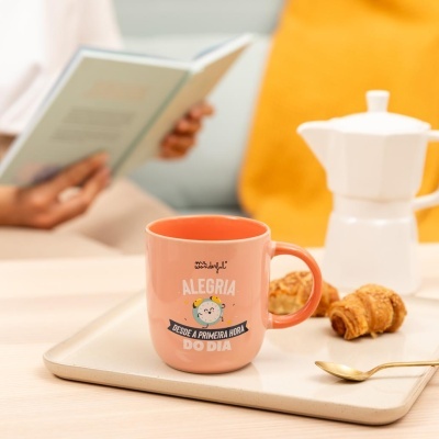 Caneca cor de salmão com texto motivacional em tabuleiro com croissants e colher dourada.
