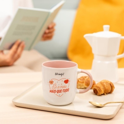 Caneca rosa e branca com texto e dois corações felizes numa bandeja com croissants e cafeteira