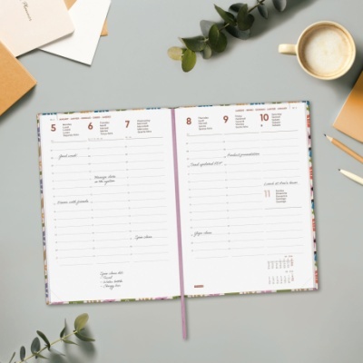 Agenda aberta com páginas de dias da semana, fita roxa, sobre mesa com plantas e chávena de café