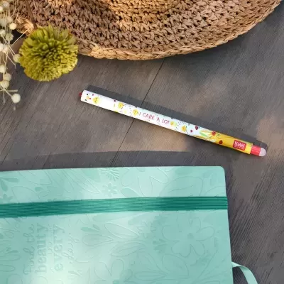 Lápis decorativo com pintainhos e texto, caderno verde e objeto de palha numa mesa de madeira