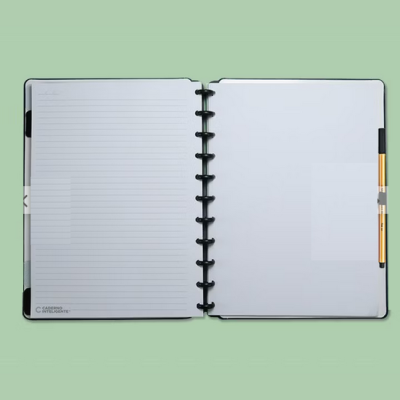 Caderno de argolas preto com folhas brancas e caneta dourada sobre fundo verde