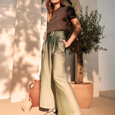 Mulher com t-shirt preta, calças largas verde claro com cinto, sapatilhas multicoloridas em ambiente areia com vasos e planta