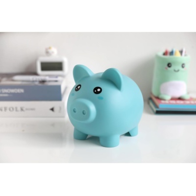 Cofre porquinho azul de plástico com olhos grandes numa mesa branca com livros e porta-lápis ao fundo